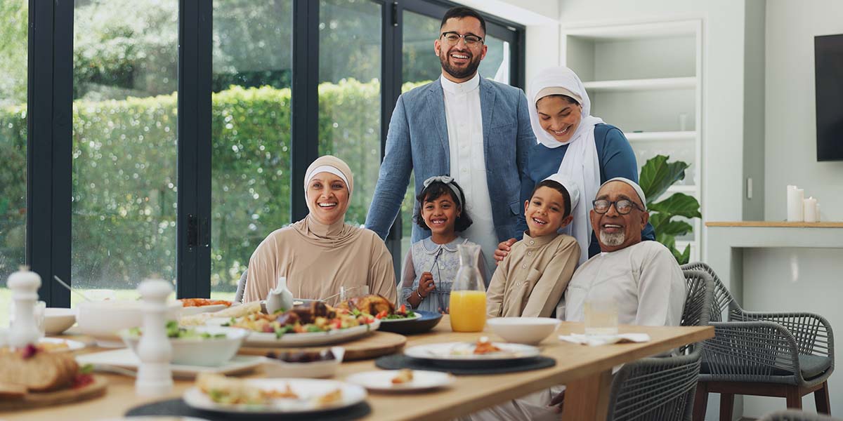 A joyful family gathers around a dining table filled with diverse dishes. The group, smiling and wearing traditional clothing, is set in a bright room with large windows.